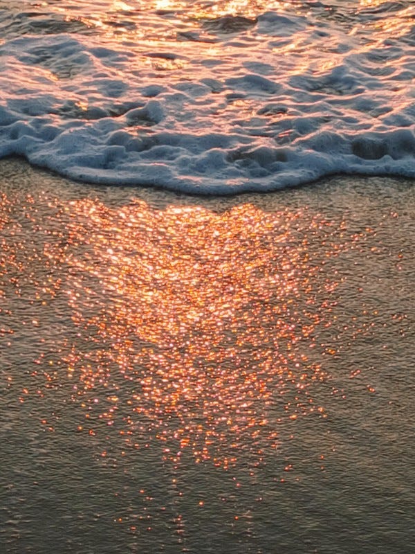 Photo en gros plan du rivage de l'océan au coucher de soleil, juste après qu'une vague se soit retirée: on voit le sable humide recouvert de petites bulles d'écume et des reflets orangés du soleil couchant avec en arrière plan l'écume bouillonante de la vague qui s'est retirée