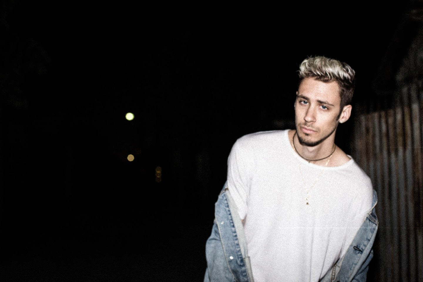  cøzybøy in a white shirt and denim jacket fall off his shoulders staring at the camera in front of a dark outdoor background.