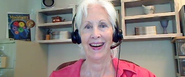 Author, Donna Blevins, smiling wide with white hair, headset, and hot pink blouse. In her office with her book’s poster, MindShift On-Demand over her right shoulder. A hanging shelf behind her with books, blue ceramic goblet, grinder, copper tea pit, and white mortar and pestle.