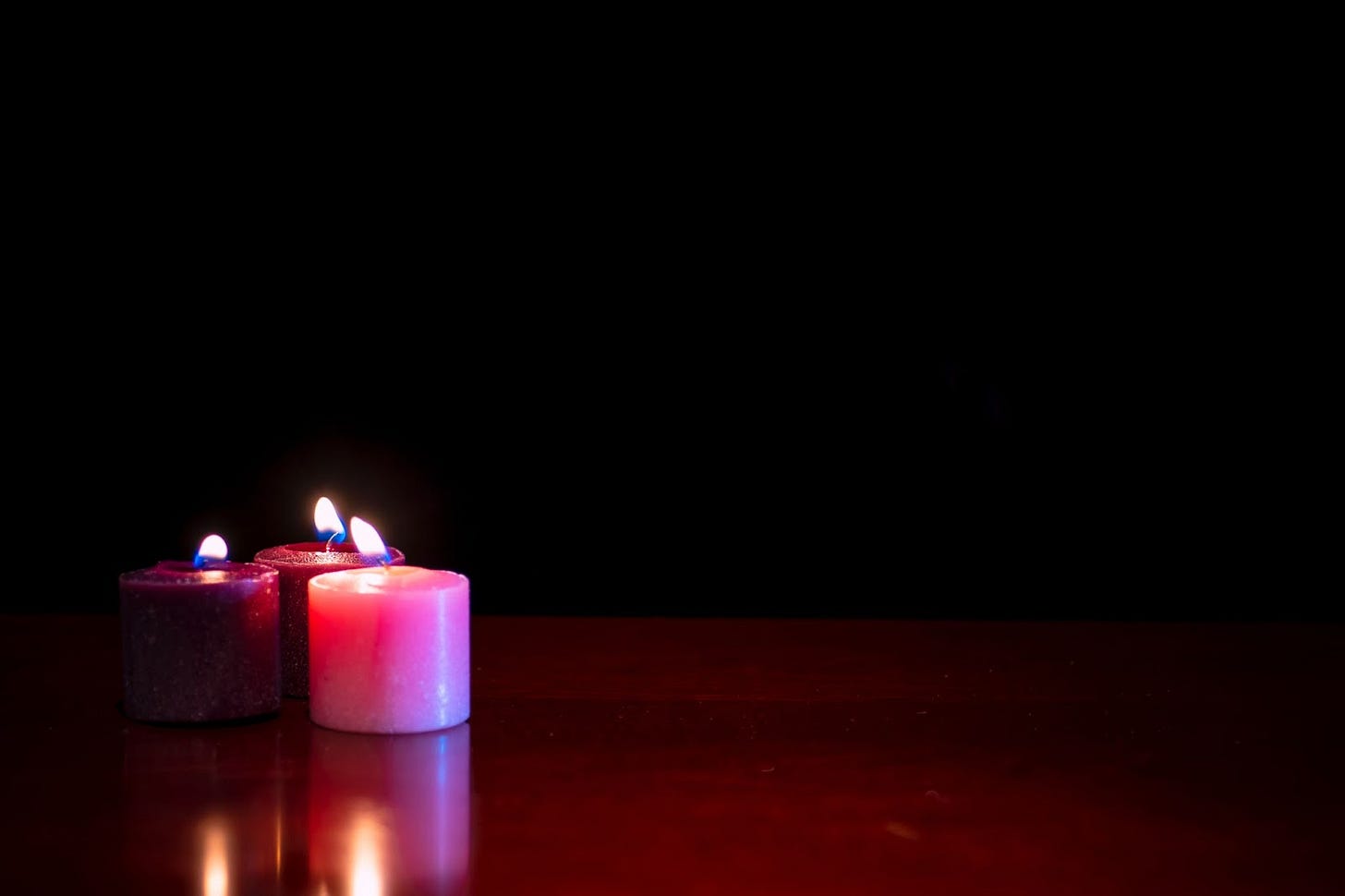Two purple and one pink candles burning on a hardwood floor with a black background. Two purple and one pink candles burning on a hardwood floor with a black background.