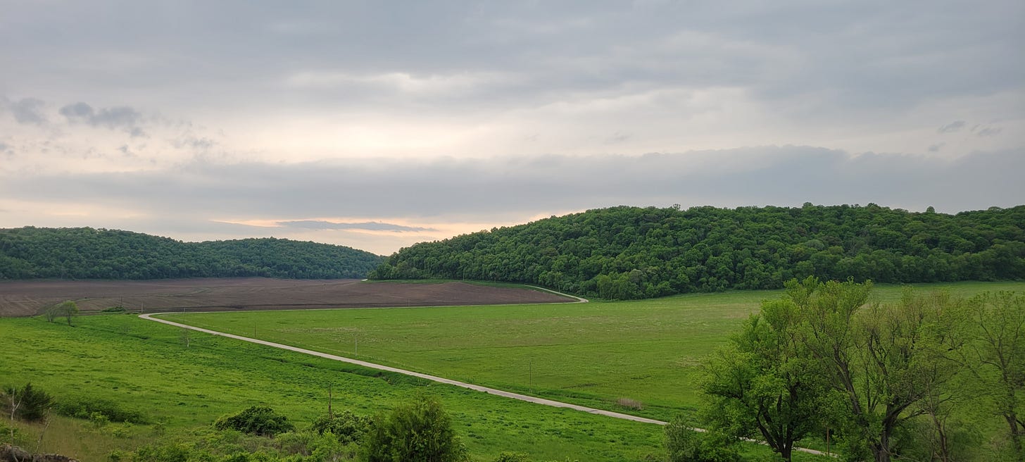  A couple of tree-covered ridges in the background, with tilled fam land, grassy fields, and a country road cutting across the fields and between the ridges. The sky is grey and cloudy.