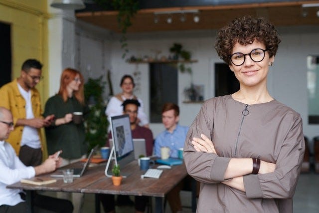person standing in the foregorund with arms crossed, smiling and looking at the camera. in the background out of focus there are 6 more people seated or standing around a wooden table. they are in a bright office space