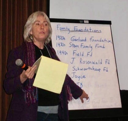 A white middle aged women standing in front of a white board, talking to an unseen audience. The white board has a list of foundations on it. Terry has silver hair and glasses and is holding a yellow legal pad.
