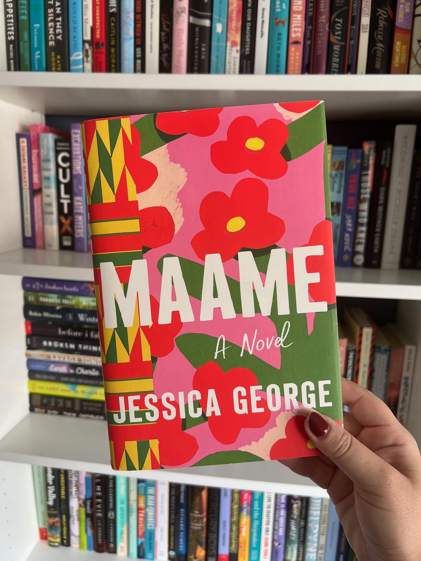 A hand holding up the book Maame in front of bookshelves. A hand holding up the book Maame in front of bookshelves.