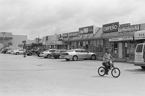 Counter-clockwise from top left: view from photographer's eye-level perspective of her feet in converse sneakers; three customers framed by two cars in adjacent parking lot at a ventanita, or coffee window, of a Cuban restaurant; portrait of young boy looking straight to camera with trees in background; image of photography lab; skyward view of sun poking through oak trees with and American flag in opposite corner; landscape view of a strip mall in suburban Miami.