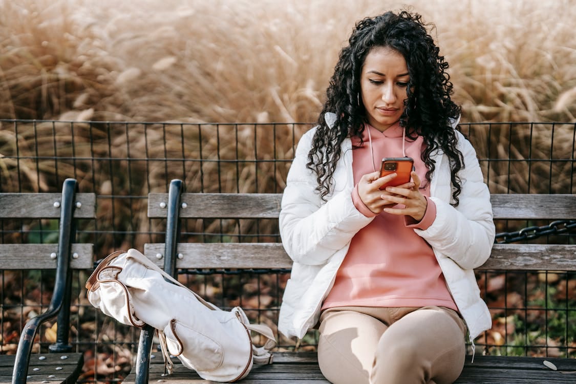 A young Latine woman with long curly dark hair sitas on a park bench as she reads her phone.