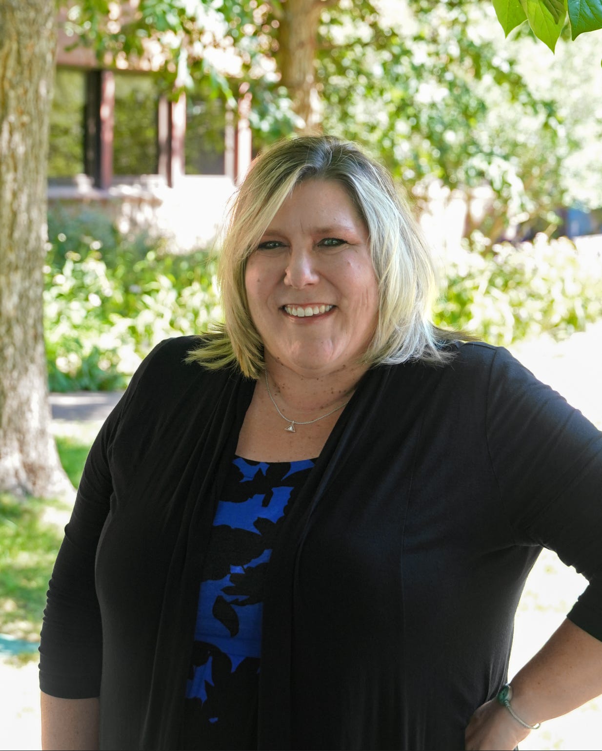 Kelly DiMartino stands outdoors in front of leafy trees and a building with large windows, smiling warmly with one hand on her hip. She is wearing a black cardigan over a blue and black patterned dress, with a necklace and bracelet. Sunlight filters through the trees, giving the scene a welcoming and professional feel.