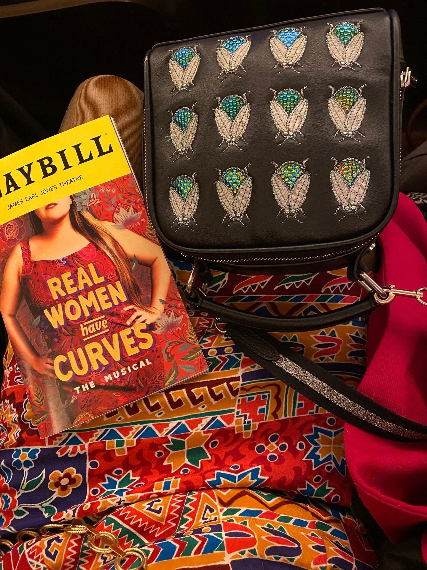 The Real Woman Have Curves playbill in Kiana's lap along with her black purse that has blue and green beetle pattern on it. The red patterned skirt coordinates with the Playbill very well.