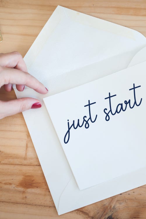 White note card and envelope with woman's hand in view. She has red short nails. The words "just start" are in navy blue script on the note card.