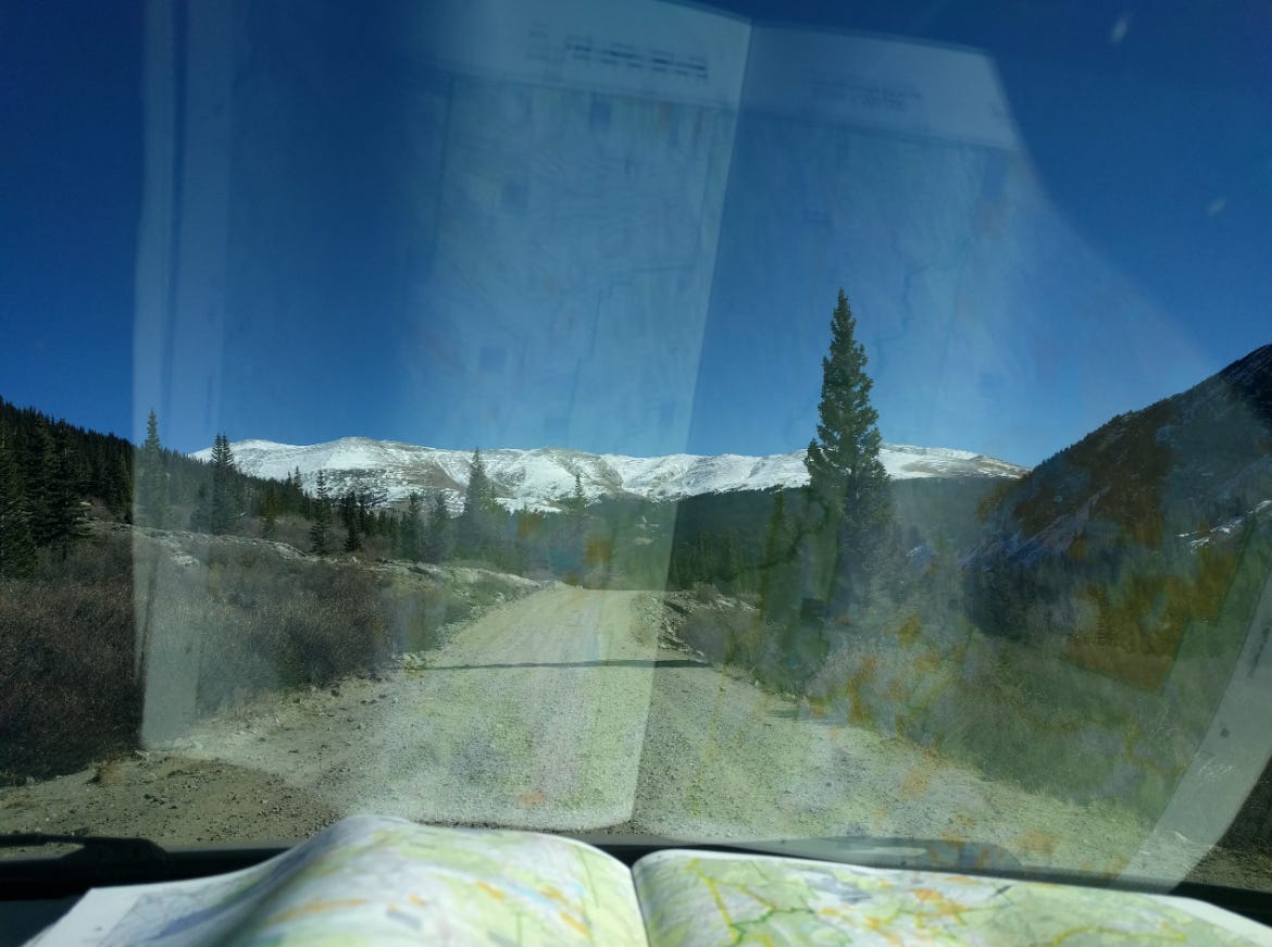 A snowcapped mountain ridge surrounded by treed ridges. A dirt road in the foreground. Reflected in a car windshield is a map of the area.