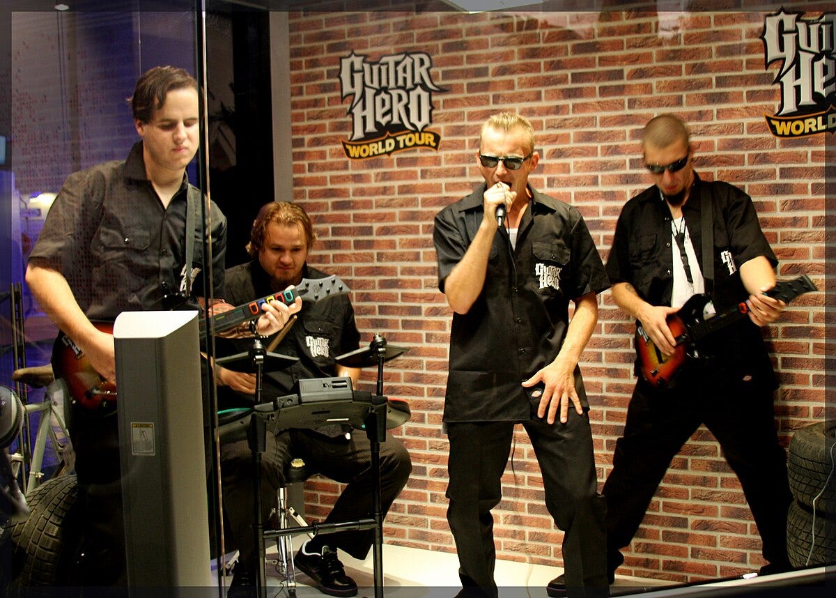 Four guys play Guitar Hero World Tour behind a glass case. One of them is wearing pit viper sunglasses and just absolutely belting it into the microphone.