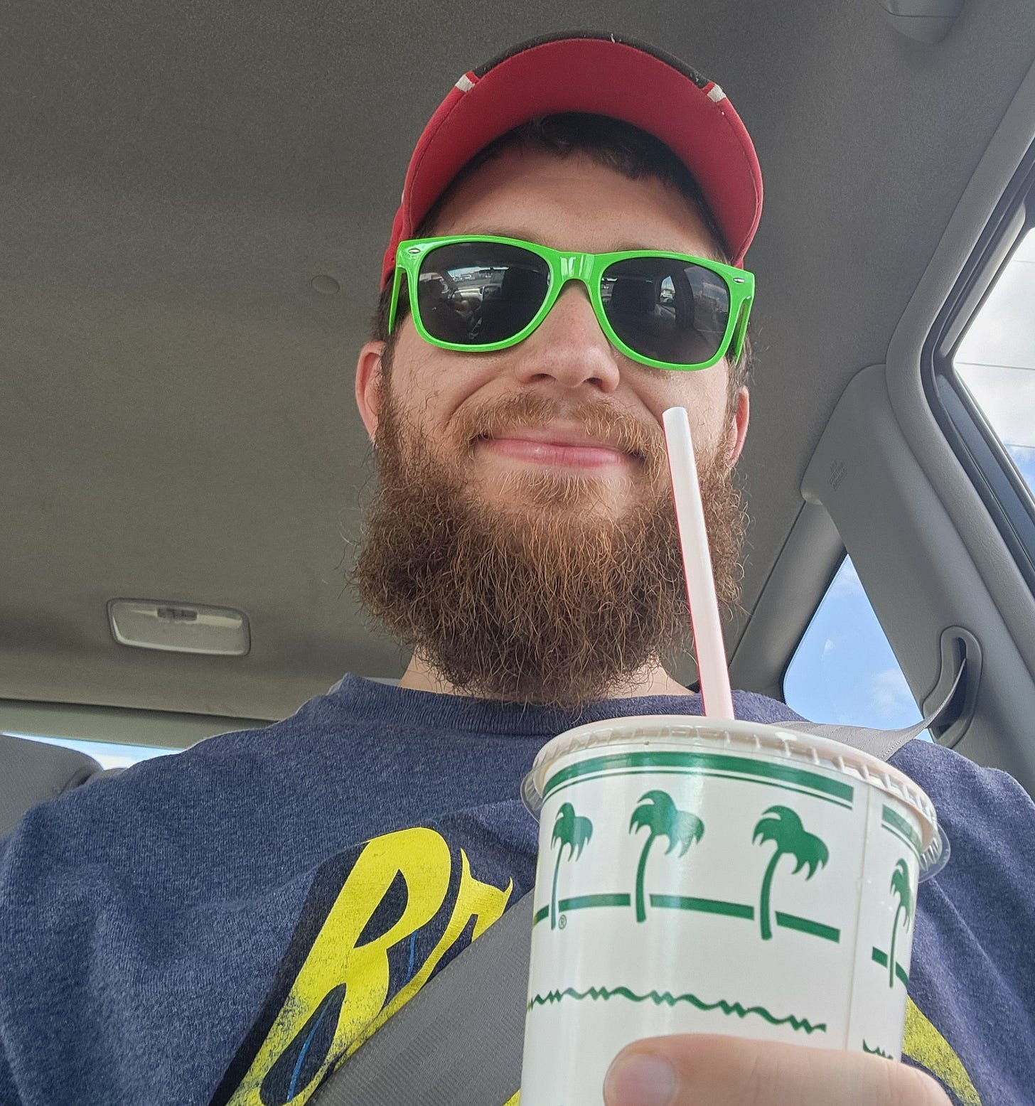 Adult male wearing a dark blue shirt reading "Batman" poses with an In-N-Out cup with green palmtrees, while sitting in a vehicle with a red hat and green sunglasses.