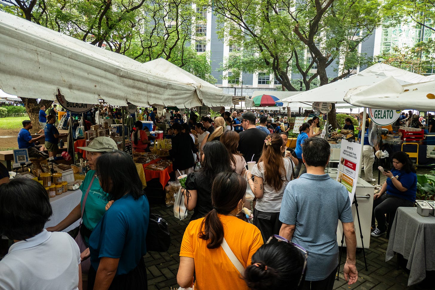 Photo of crowded farmer's market in Makati, Manila, Philippines. Beautiful large trees provide shade for the stalls of the market, each stall with a white pop up tent and tables with their goods. The aisle between two rows of vendors is packed with people shopping and walking.