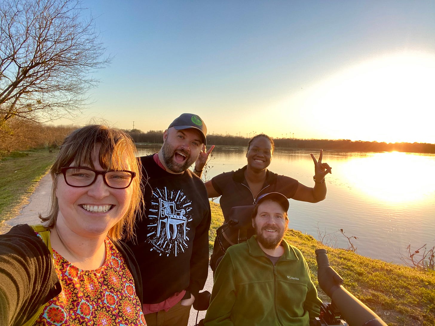 Four smiling adult friends in their thirties pose for a selfie at sunset. At left, Beth holds the camera with her outstretched hand. She wears glasses and a warm-colored top with abstract designs. Tyler sports a beard and a ball cap and a black sweater with a print of a white, wooden chair on the front. Eric also has a beard and ball cap and wears a green fleece jacket. Mounted to the tray of his motorized wheelchair is a large spotting scope. Standing behind Eric, Kira wears a black top and makes peace signs with her fingers. An expansive pond fills the background while a large, leafless tree looms over Beth. The sun is bright as it sinks toward the horizon. Four smiling adult friends in their thirties pose for a selfie at sunset. At left, Beth holds the camera with her outstretched hand. She wears glasses and a warm-colored top with abstract designs. Tyler sports a beard and a ball cap and a black sweater with a print of a white, wooden chair on the front. Eric also has a beard and ball cap and wears a green fleece jacket. Mounted to the tray of his motorized wheelchair is a large spotting scope. Standing behind Eric, Kira wears a black top and makes peace signs with her fingers. An expansive pond fills the background while a large, leafless tree looms over Beth. The sun is bright as it sinks toward the horizon.