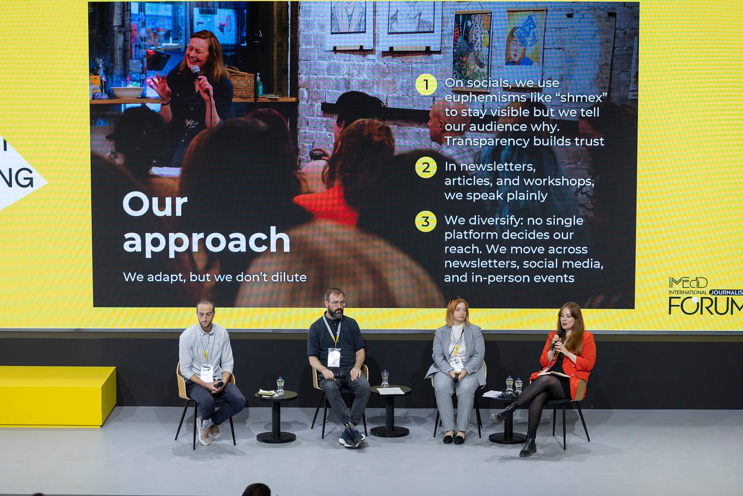 A wide shot of a journalism panel at the iMEdD International Journalism Forum in Athens. Four speakers sit on stage in front of a large yellow screen that reads “Our approach – We adapt, but we don’t dilute.” The screen lists Pillow Talk Scotland’s strategies for navigating online censorship, while founder Iris Pase, wearing a red blazer, speaks into a microphone.