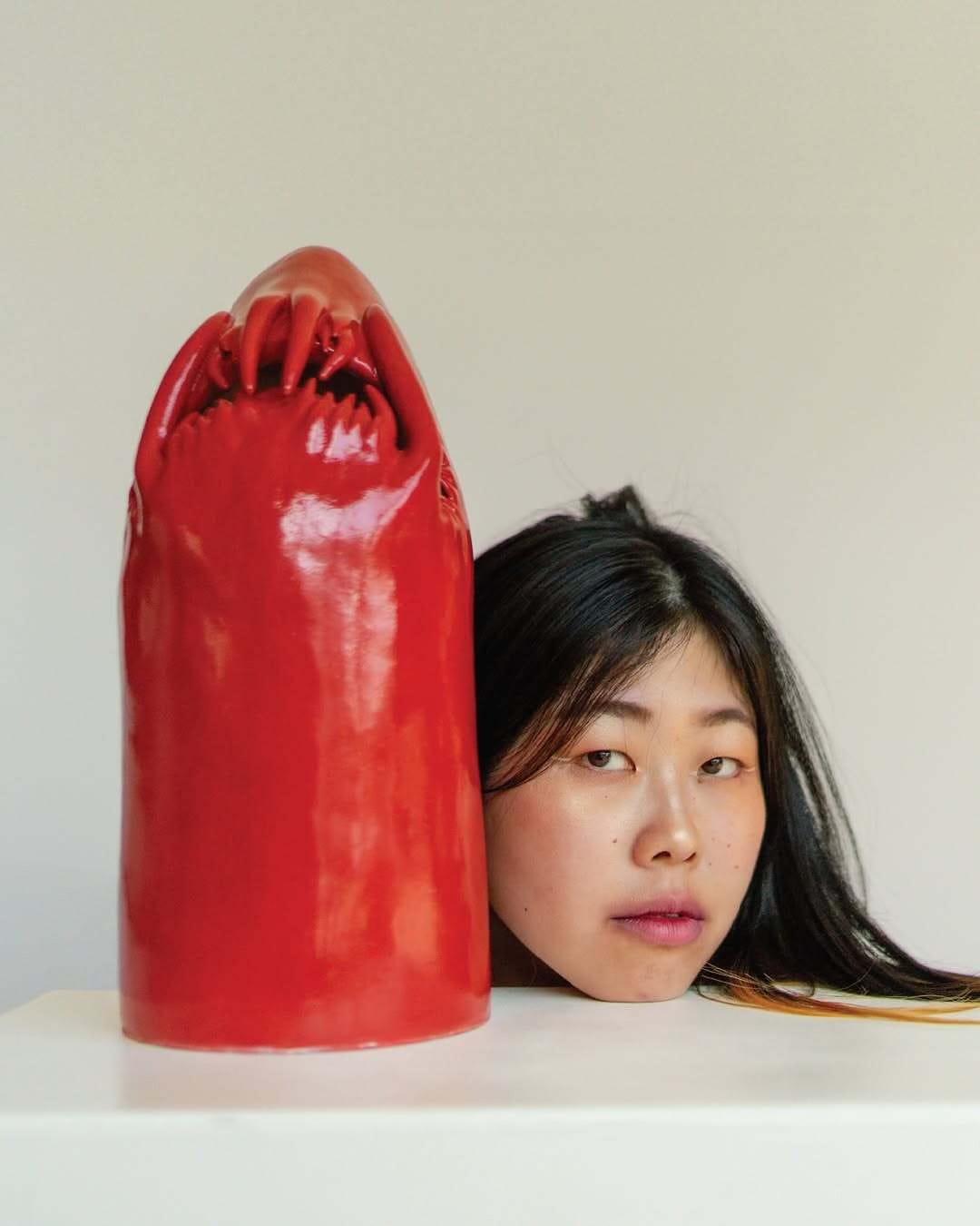 Portrait of artist Eri Maeda with her face resting beside a tall, glossy red ceramic sculpture shaped like a surreal claw. The work blurs lines between body, tool, and protection. Artist Eri Maeda resting her face beside a tall red ceramic creature with sculpted fingers on top, highlighting her surreal hand built approach to contemporary ceramics.