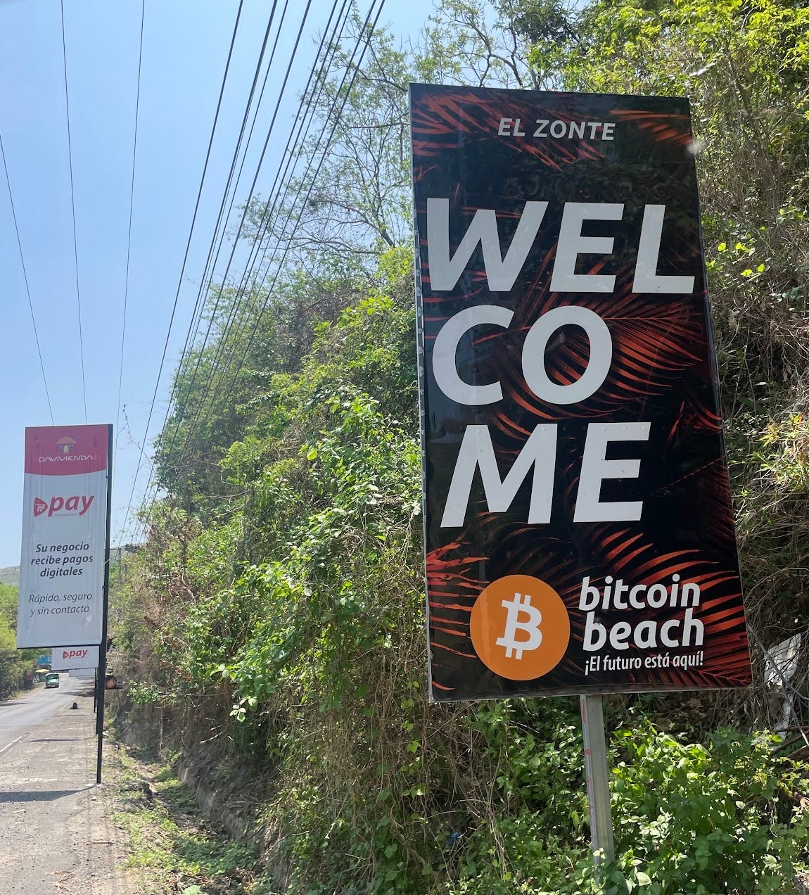 A roadside sign says, in English, "El Zonte βΒ WELCOME βΒ Bitcoin Beach," and below in Spanish, "El futuro esta aqui!" A roadside sign says, in English, "El Zonte βΒ WELCOME βΒ Bitcoin Beach," and below in Spanish, "El futuro esta aqui!"
