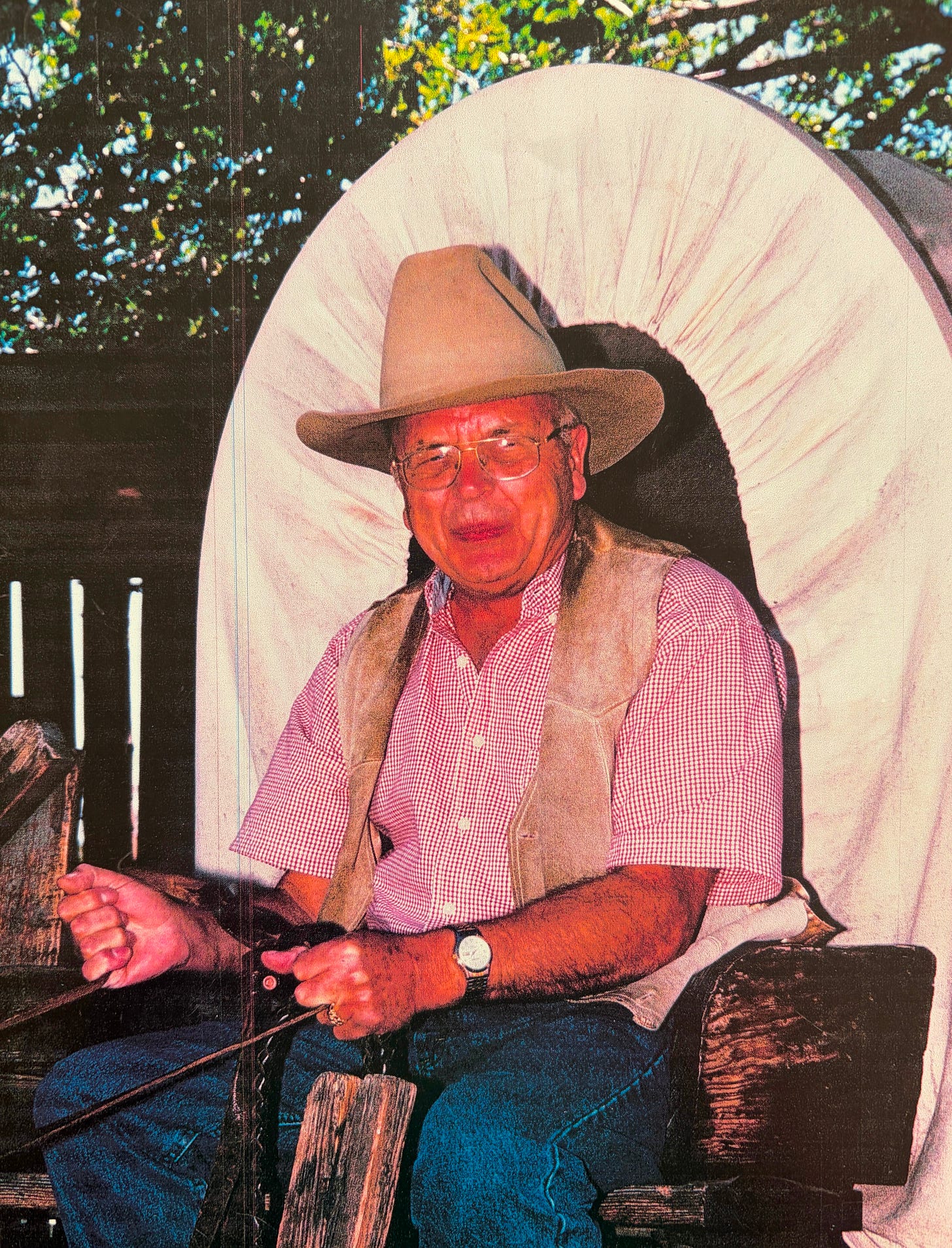 Bespectacled Grandfather wearing a beige cowboy hat, and vest as well as red and white checked shirt with a caravan behind him and reins in his hands.