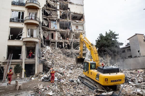 A multistory building with an exterior wall sheared off, with debris hanging from it and in a tall mound in front of it. A yellow front- loader is poised to dig into the debris pile. A multistory building with an exterior wall sheared off, with debris hanging from it and in a tall mound in front of it. A yellow front- loader is poised to dig into the debris pile.
