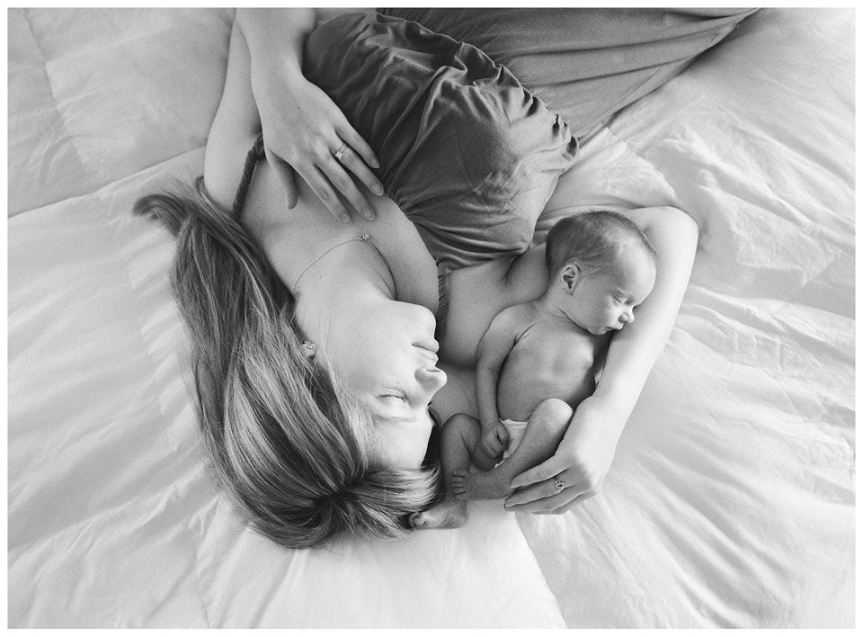 A black and white photo of a woman with blonde hair laying on her bed. Her arm is up and her newborn baby is nestled in the space. Their profiles look identify.