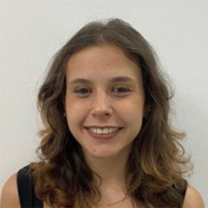 Jovem de cabelos castanhos ondulados e sorriso discreto. Veste blusa preta sem mangas e colar com pingente dourado. Jovem de cabelos castanhos ondulados e sorriso discreto. Veste blusa preta sem mangas e colar com pingente dourado.