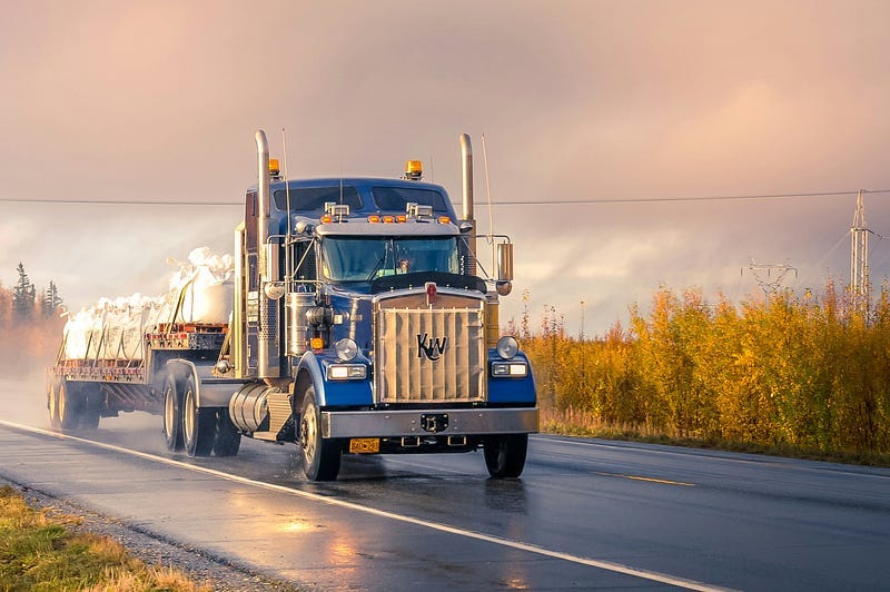 Blue tractor trailer hauling a load down a two-lane highway, storms behind and sun ahead.