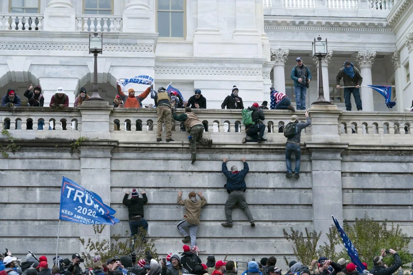 Individuals climbing a wall outside the U.S. Capitol with others standing on a balcony watching and holding flags.