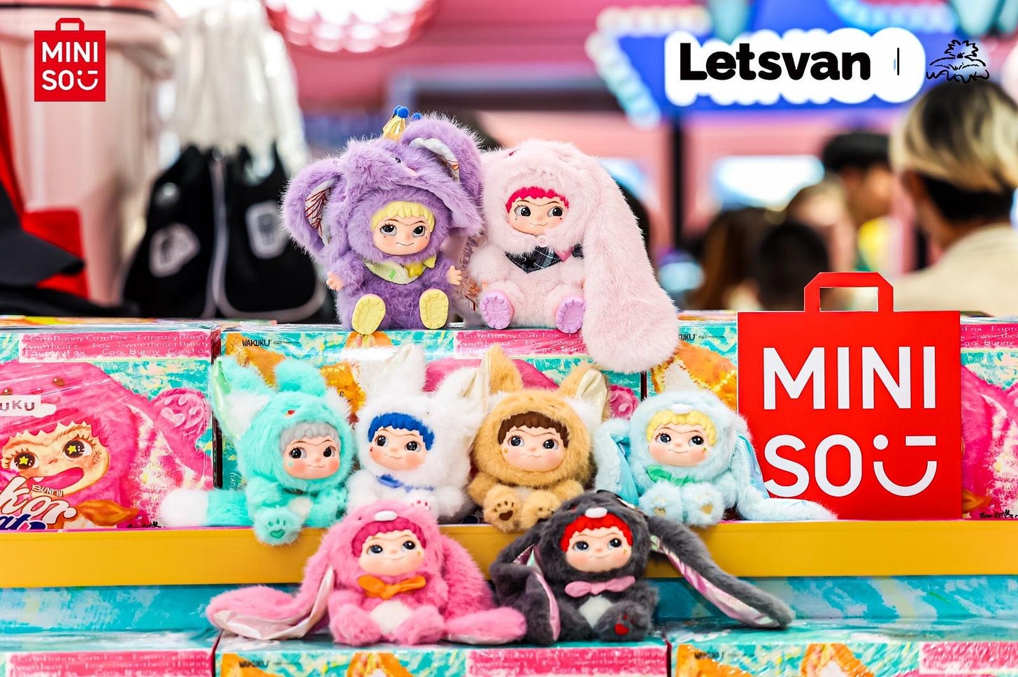 Eight miniature Wakuku plush toys in multicolor outfits arranged on product boxes beside signature red Miniso bag.