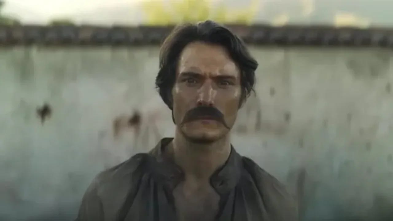 homem  branco de cabelos pretos e vasto bigode, magro, no centro da foto, olha fixamente para a câmera. atrás um muro branco desfocado.