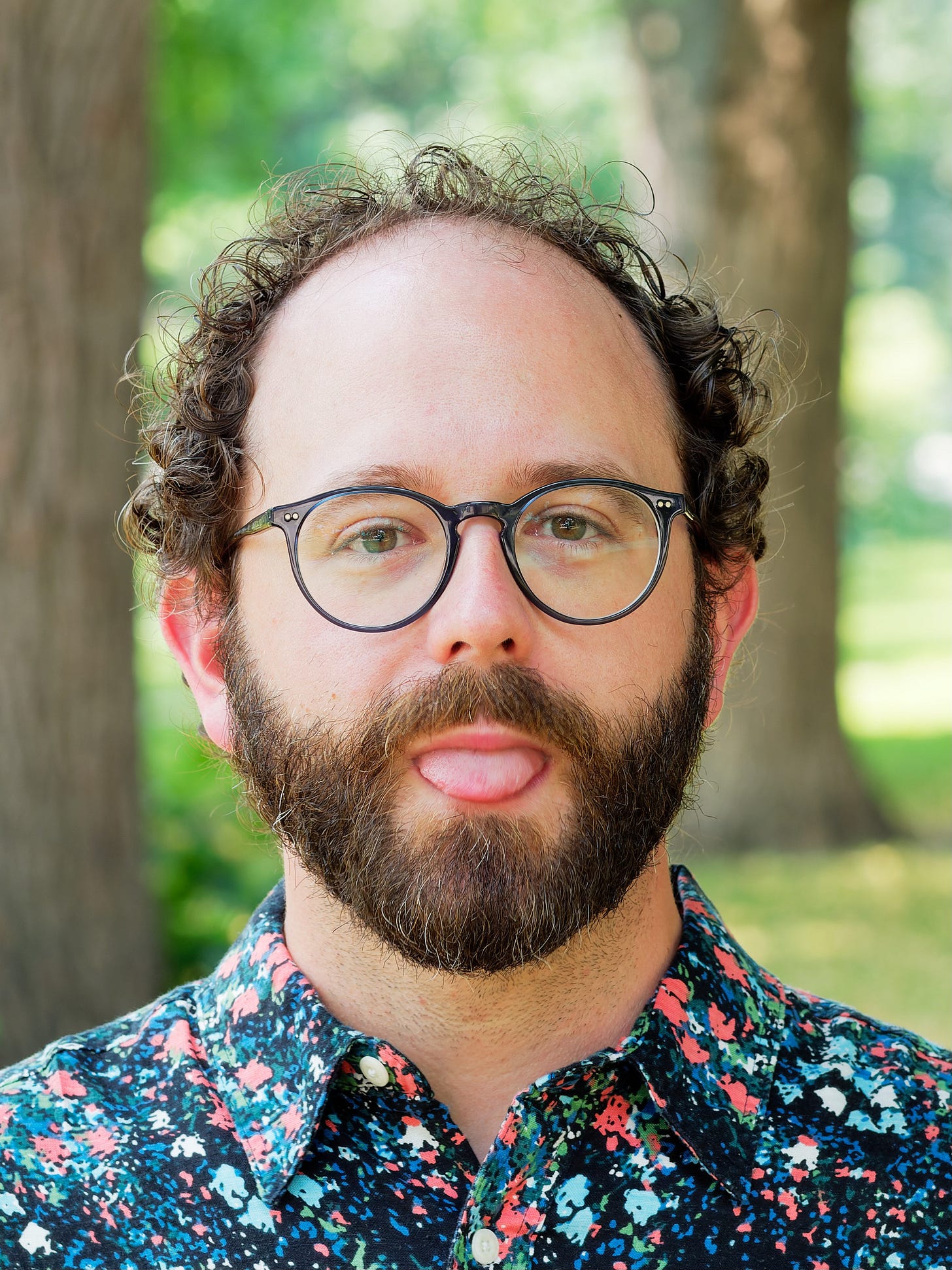photo of a silly man with curly hair and glasses and beard standing in a park sticking out his tongue wearing a multicoloured shirt photo of a silly man with curly hair and glasses and beard standing in a park sticking out his tongue wearing a multicoloured shirt