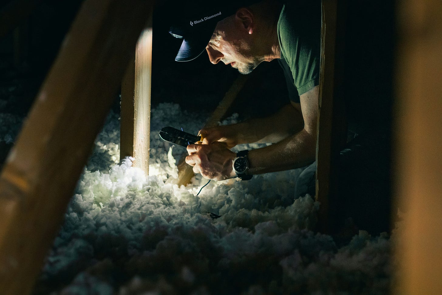 A man crawling through attic insulation shines a flashlight while working with tools in the attic. A man crawling through attic insulation shines a flashlight while working with tools in the attic.