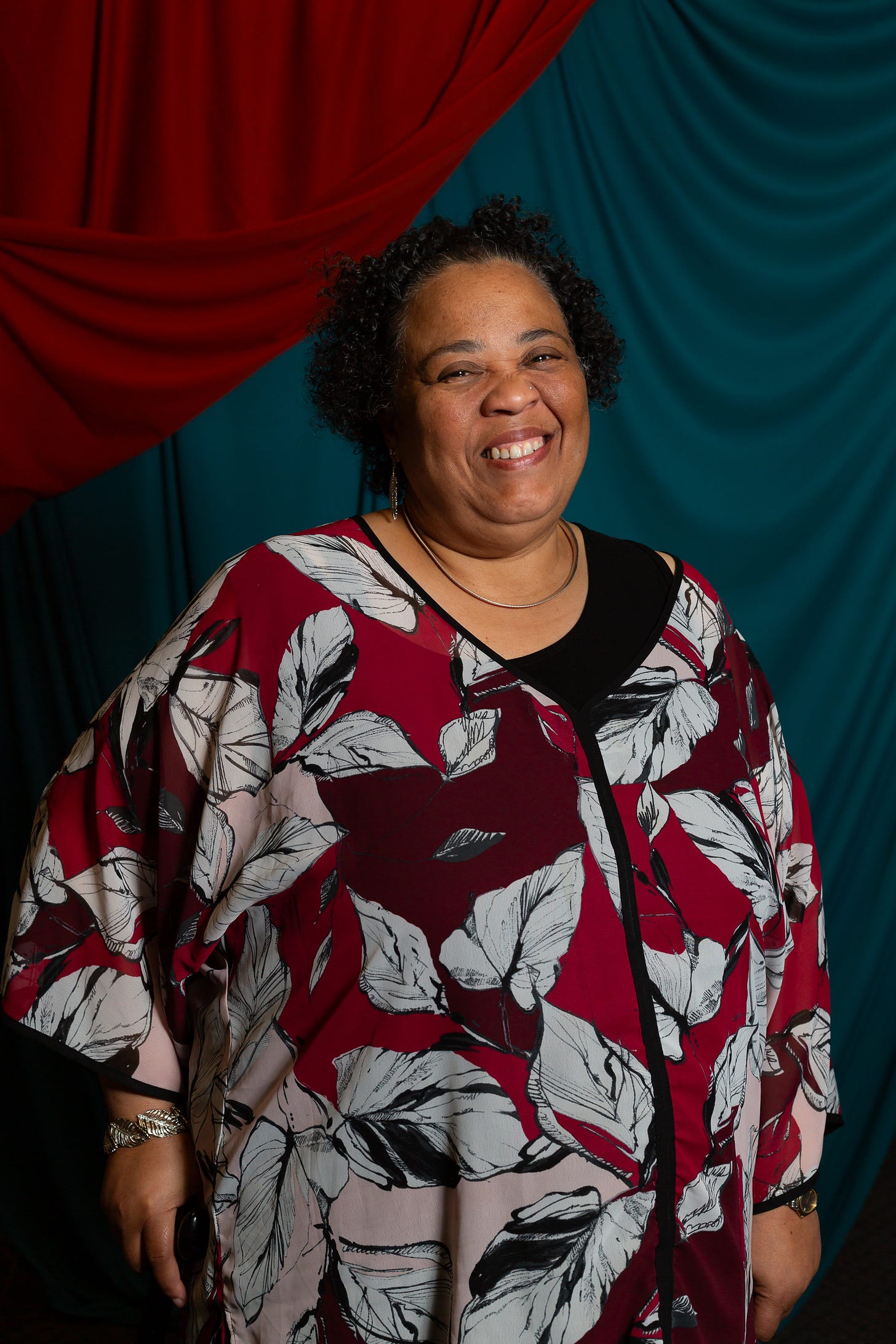 Portrait of a dark skinned woman with dark short curly hair and brown eyes. She is smiling at the camera. She is wearing silver hoops, a silver necklace and silver leaf bracelet. She is wearing a flowy, floral red, white and black top. She holds a cane in her right hand. 