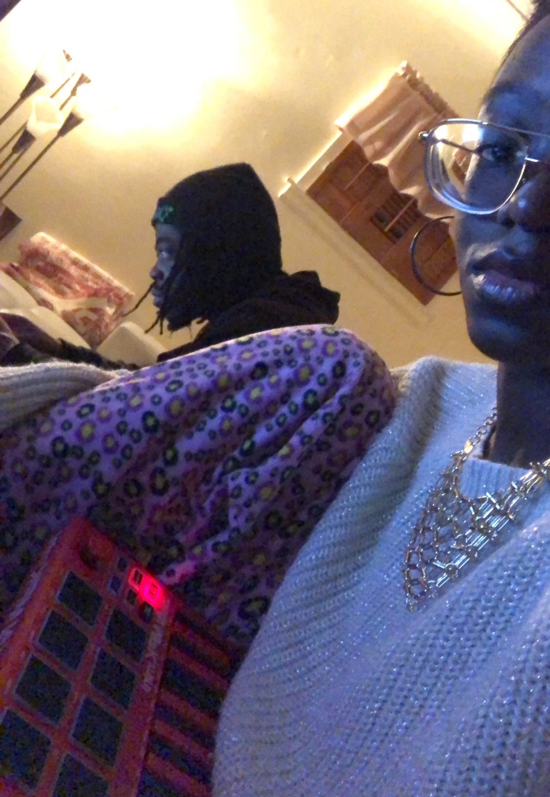 dark-skinned Black girl wearing a cream sweater with gold threading, a gold necklace and pink leopord print pajama pants sits with a red MPC machine on her lap. In the background, a brown-skinned Black man with locs and a black beanie works on a laptop.