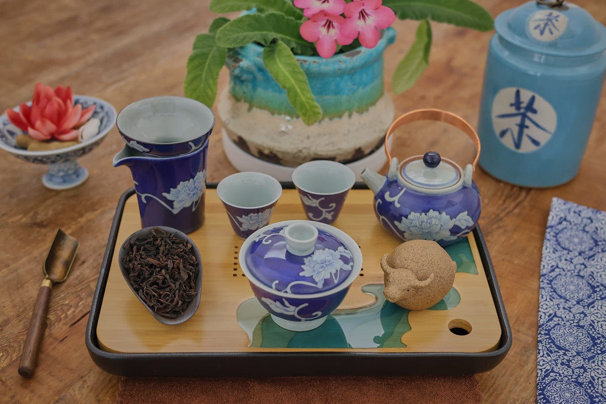 a traditional gong fu tea ware preparation with a flowering plant in the background, napkin, and canister in the background a traditional gong fu tea ware preparation with a flowering plant in the background, napkin, and canister in the background
