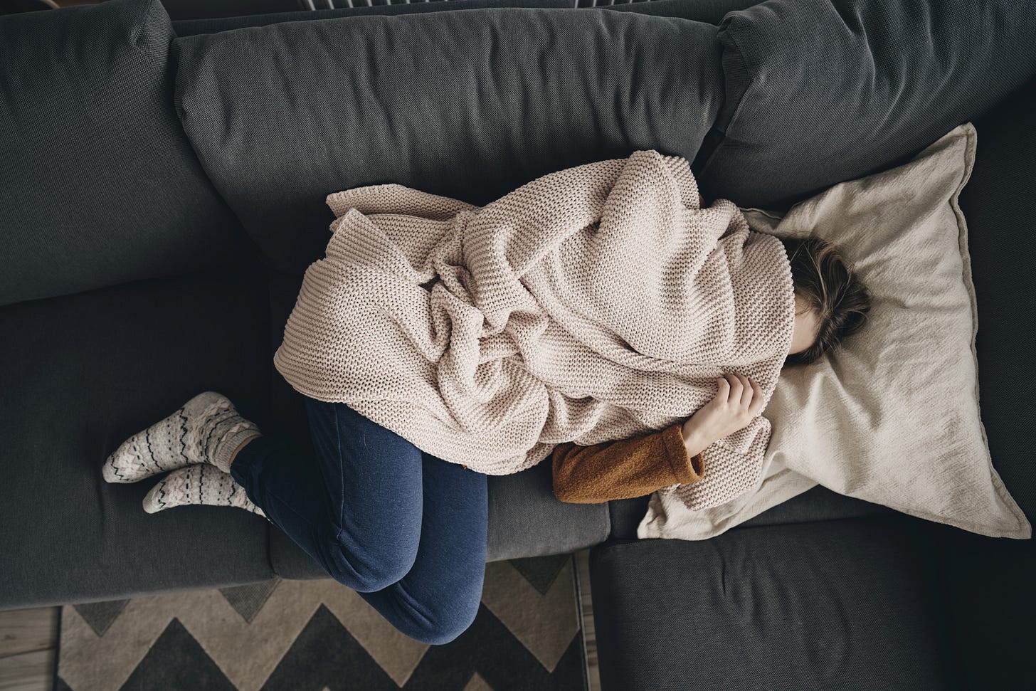 Person lying on a sofa, face and half their body hidden under a blanket. 