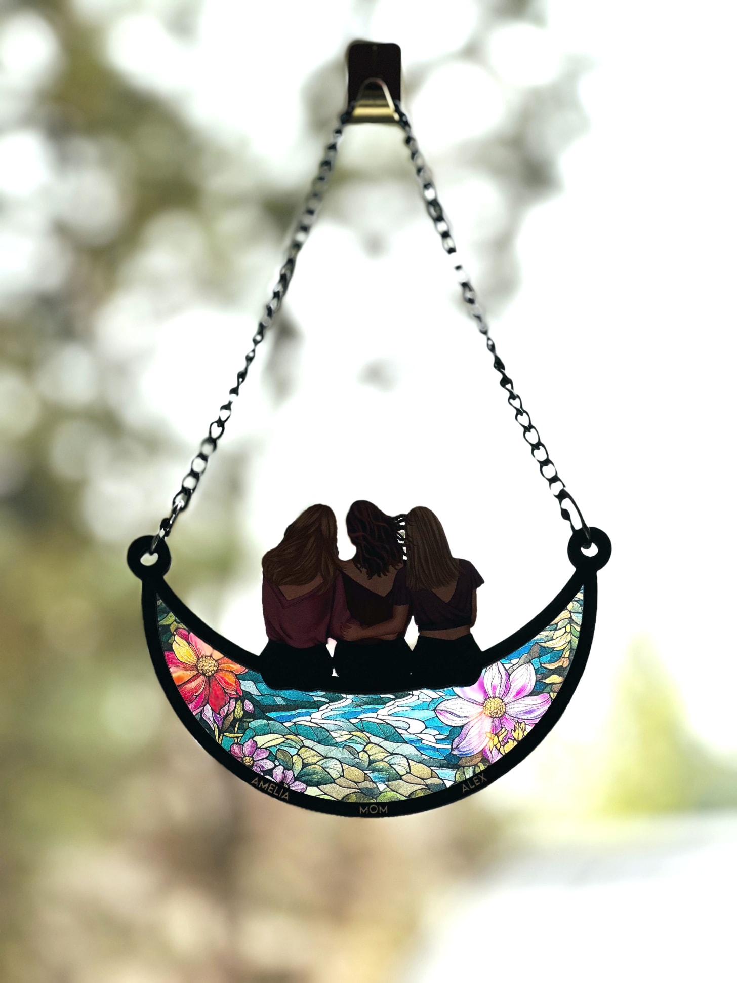 A stained glass suncatcher hanging on a window, depicting three women sitting on floral stained glass
