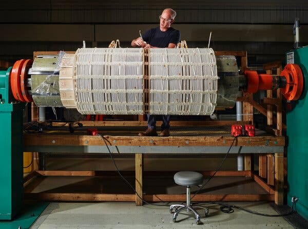A person working at a large cylindrical piece of equipment that is wrapped in coils. A person working at a large cylindrical piece of equipment that is wrapped in coils.