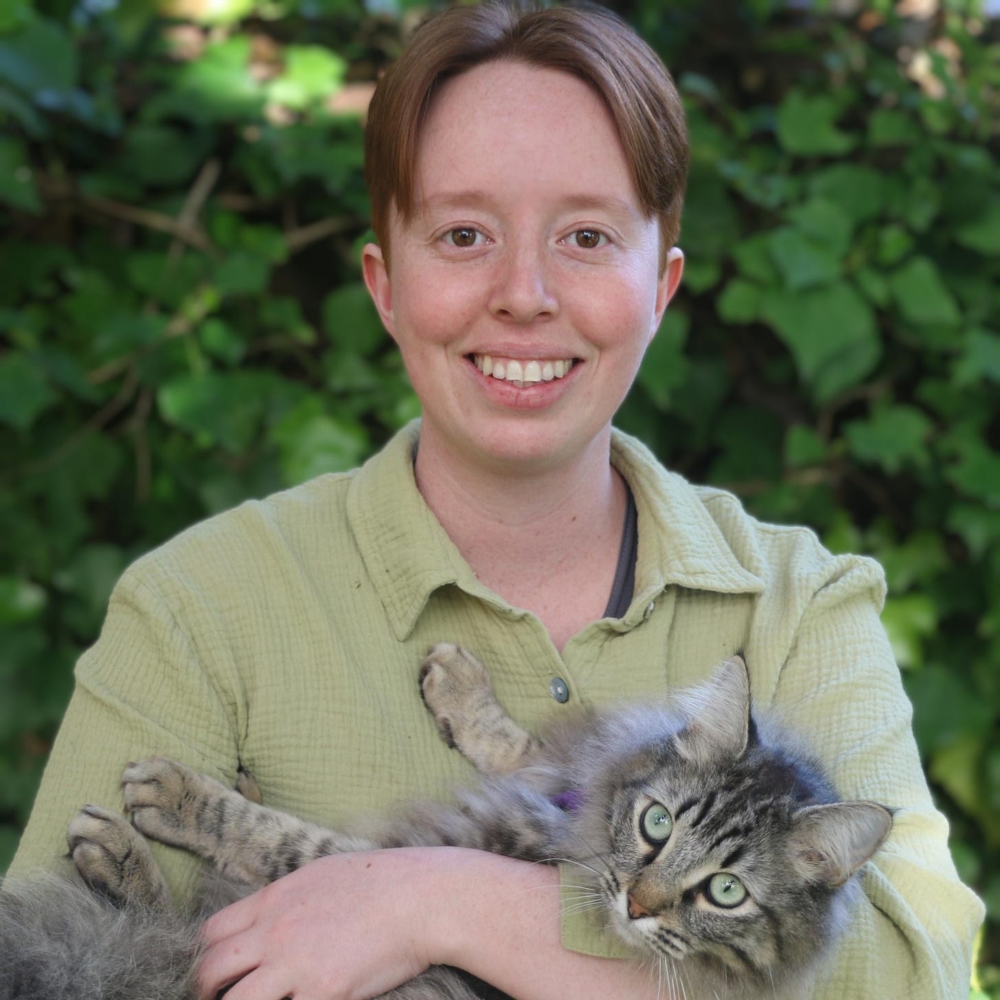 A picture of Cay Macres standing in front of a hedge and holding a fluffy grey cat A picture of Cay Macres standing in front of a hedge and holding a fluffy grey cat