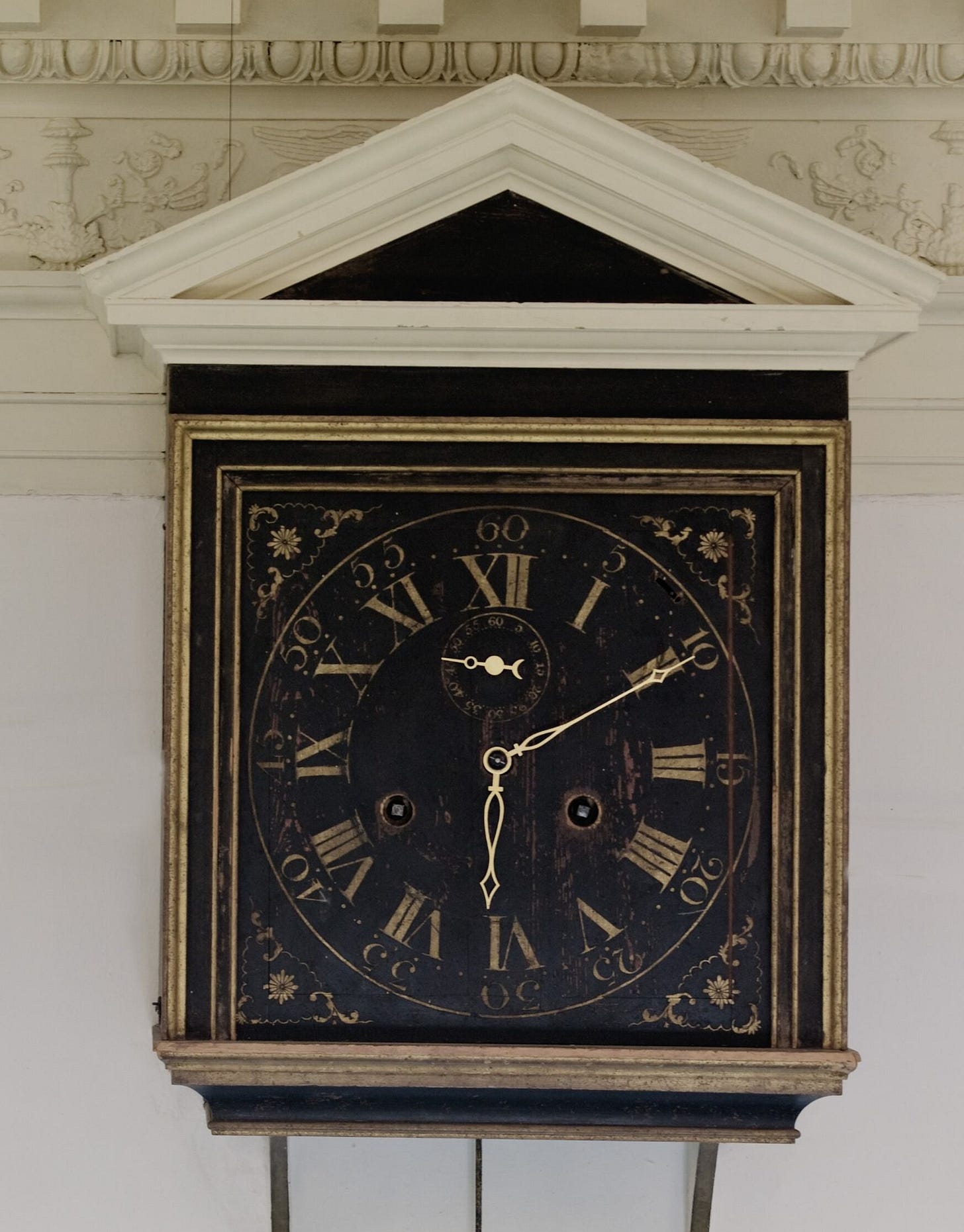 A direct shot of the interior Great Clock's interior face,  a square, painted mahogany case and separate triangular, painted pediment and three pierced hands which mark the hours in gold-colored Roman numerals, and minutes and seconds in gold-colored Arabic numerals.