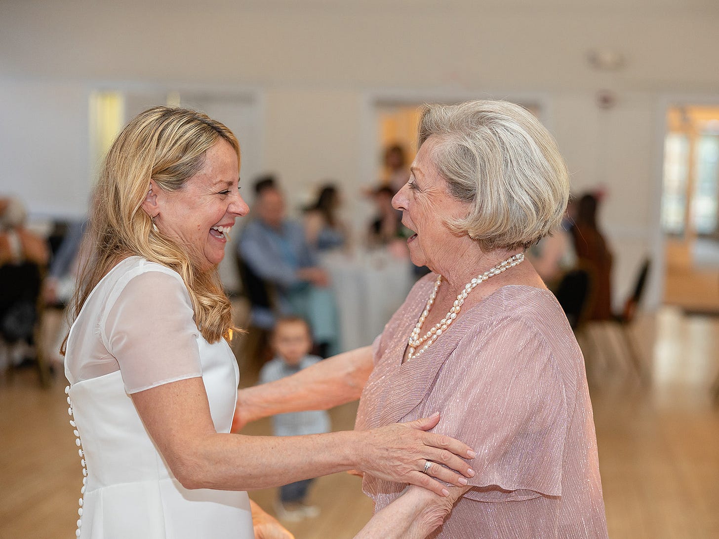 A blonde bride in a widding gown smiles and embraces her mother who wears a pink dress and pearls