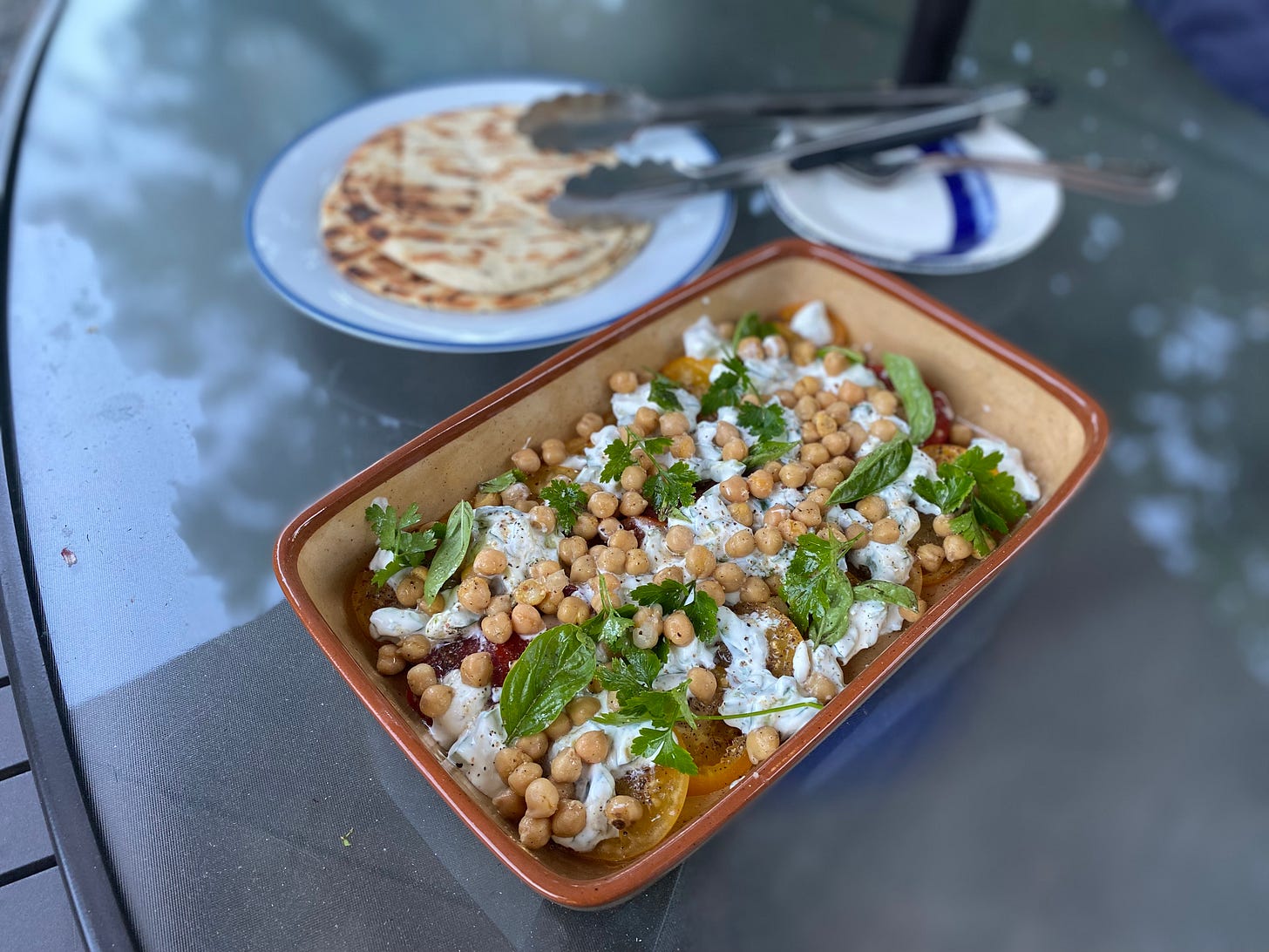 Yellow and red tomato slices arranged in a rectangular stoneware dish, with a cucumber and yogurt sauce dolloped over them. Chickpeas, parsley, and basil leaves are scattered over the top. On a large white plate are two grilled flatbread rounds, with a pair of tongs next to them. The tongs rest partially on a stack of two blue and white side plates with two forks on top of them.