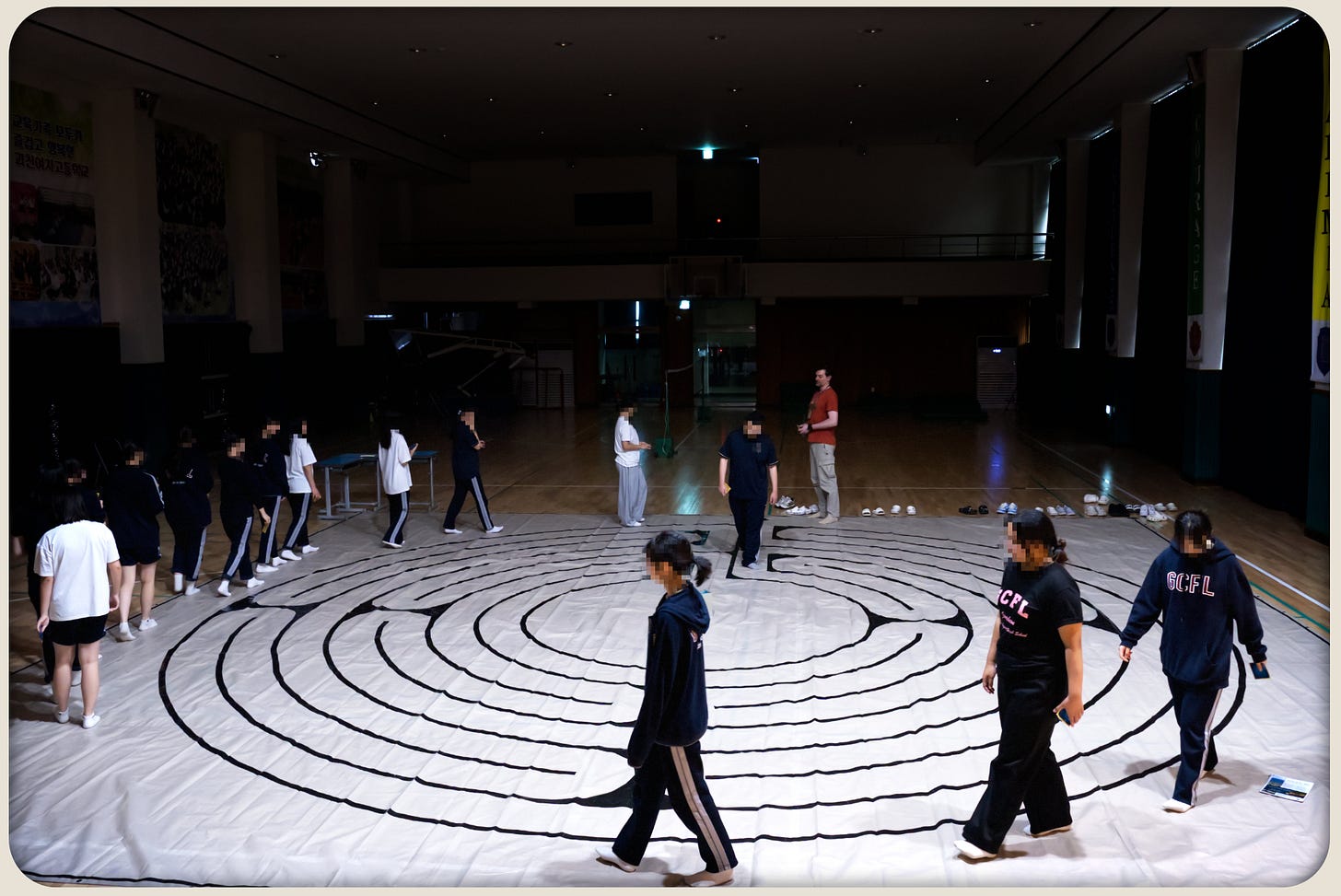 Students walk around the outer edge of a large taped labyrinth on a tarp in a dim school auditorium.