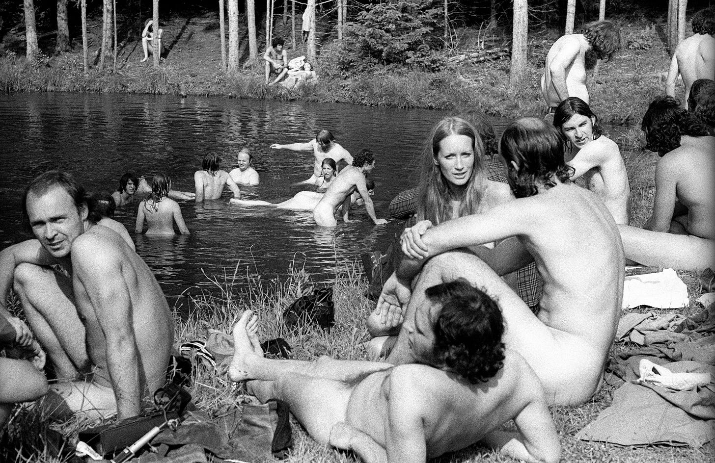 A 1970 black and white photograph of nudists bathing in a river and sitting on the grassy banks. A 1970 black and white photograph of nudists bathing in a river and sitting on the grassy banks.