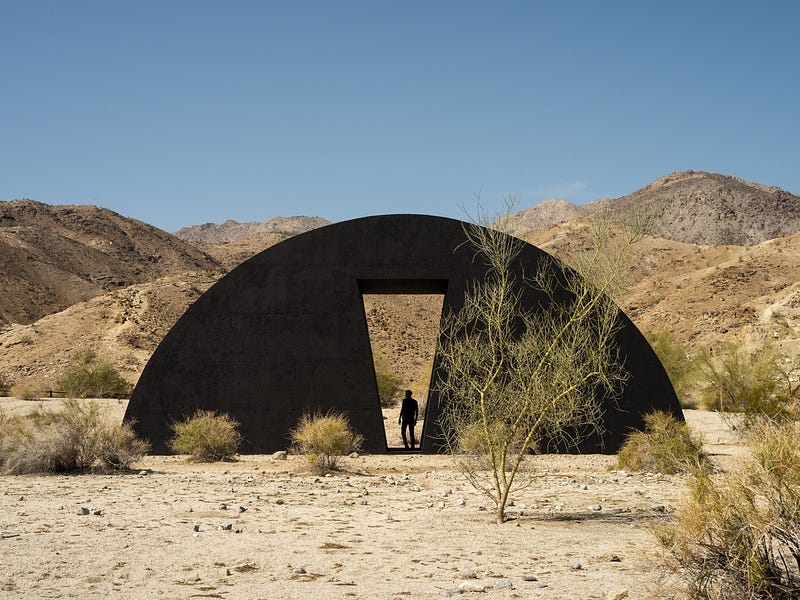 A giant, half-moon, black sculpture sits in the desert landscape. Behind it are sandy hillsides. At its center is an angular cutout, a doorway with the silhouette of a person standing inside. A giant, half-moon, black sculpture sits in the desert landscape. Behind it are sandy hillsides. At its center is an angular cutout, a doorway with the silhouette of a person standing inside.