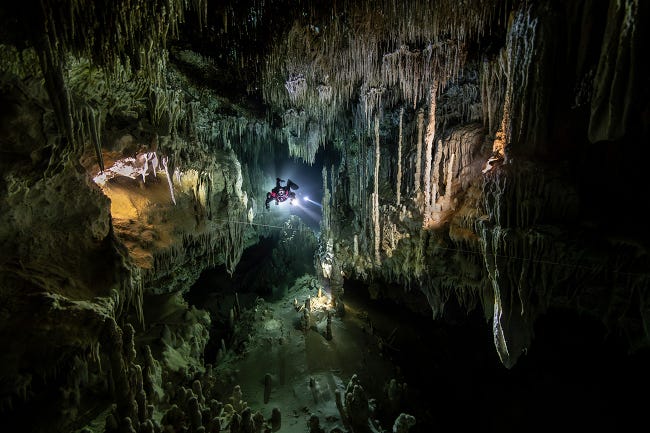Une immense caverne sous-marine où stalactites et stalgmites dessinent des formes étranges, un plongeur semble perdu en son milieu.
