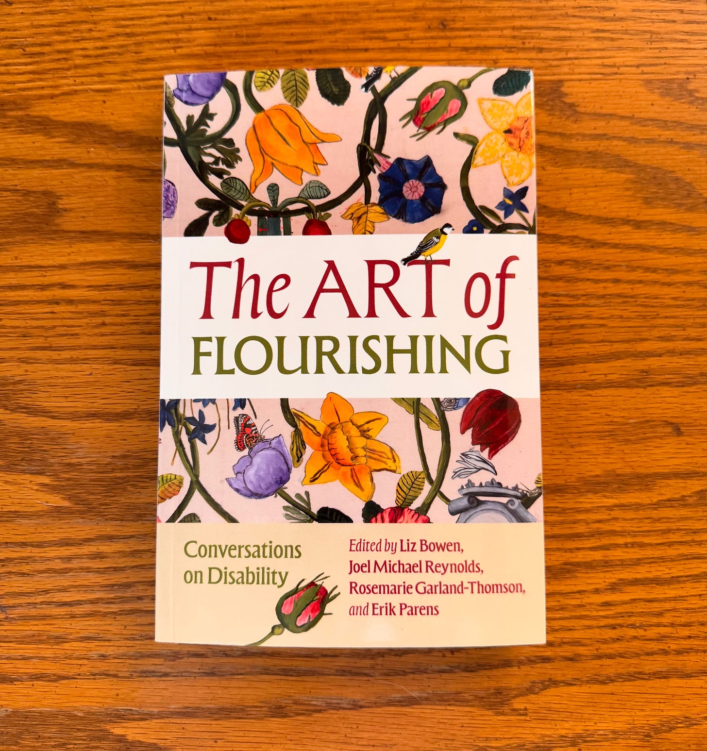 Paperback edition of THE ART OF FLOURISHING: CONVERSATIONS ON DISABILITY sits upon a light wooden table. The cover features colorful intertwined flowers behind the title, alongside small birds and butterflies.