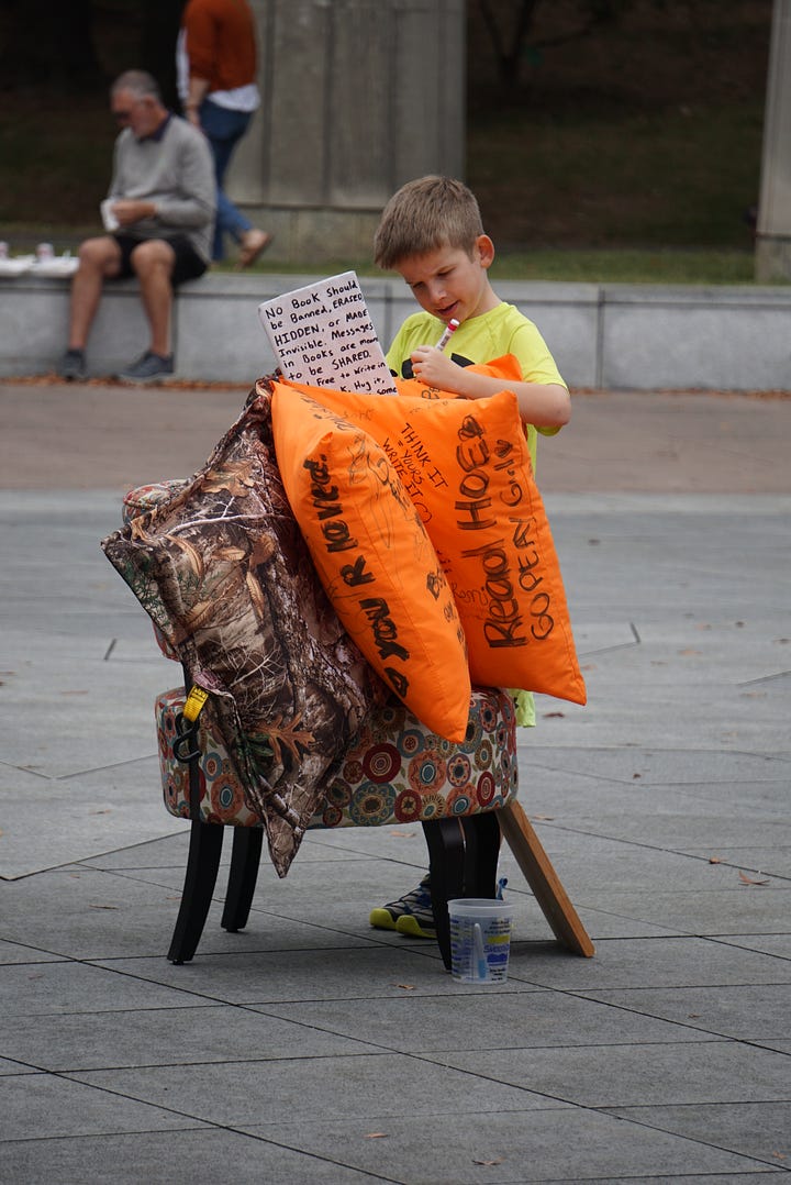 A woman holding a white bag examines a brightly colored painted giant book. A child uses a marker to add to a book made out of pillows and furniture. A group of people examines a colorful giant book with painted text and images. A giant, brightly colored book stands in front of a building with columns.