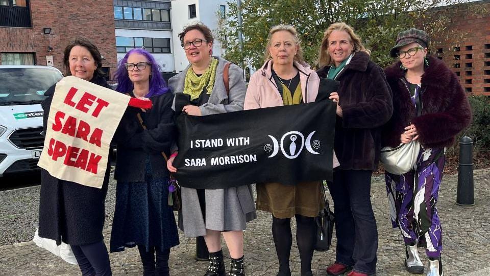 A group of women standing next to each other. There are six of them wearing a range of different coloured clothing. From left to right a woman with long brown hair, a lady with long purple hair and dark glasses. a women with short brown hair and square glasses, then there is a woman with long blonde hair and next to her is a women with blonde hair and a lady wearing a hat and green glasses. A group of women standing next to each other. There are six of them wearing a range of different coloured clothing. From left to right a woman with long brown hair, a lady with long purple hair and dark glasses. a women with short brown hair and square glasses, then there is a woman with long blonde hair and next to her is a women with blonde hair and a lady wearing a hat and green glasses.