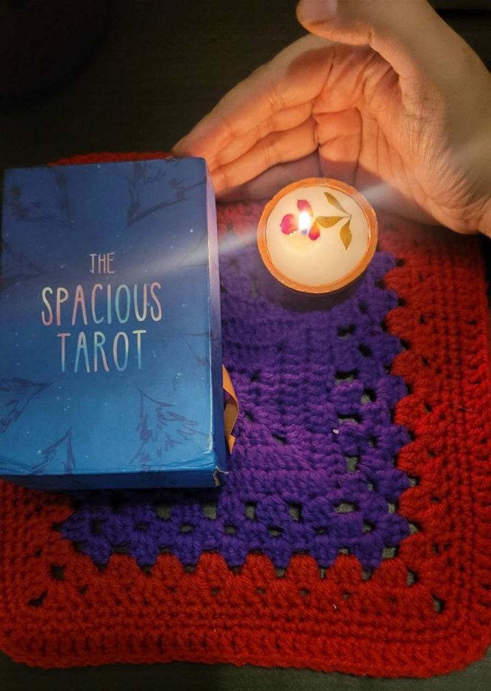 A crocheted cloth in red and blue with a tarot deck and a small candle on top. My hand on the right side is cupping the candle's flame 