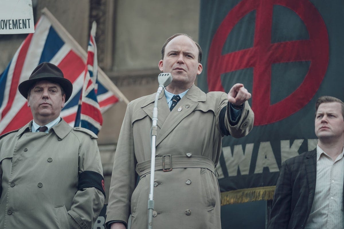 Rory Kinnear wearing a mid-20th century trenchcoat in character as Colin Jordan in BBC series Ridley Road. He is addressing a public meeting, pointing as he speaks, and stands in front of a black flag with a red cross on, to suggest fascist iconography